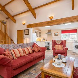 A cozy open-plan living space with a red sofa, armchair, and wooden coffee table set for tea. The room features exposed beams, a staircase, and a bright kitchen in the background.