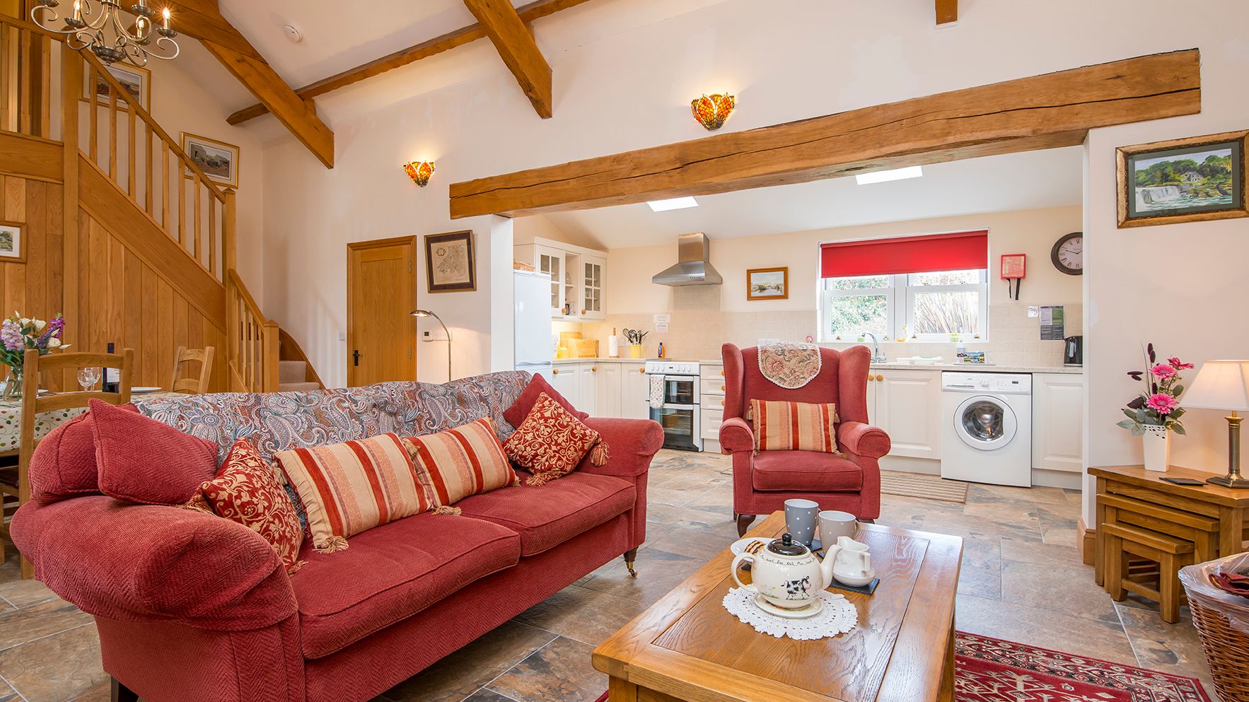 A cozy open-plan living space with a red sofa, armchair, and wooden coffee table set for tea. The room features exposed beams, a staircase, and a bright kitchen in the background.