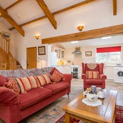 A cozy open-plan living space with a red sofa, armchair, and wooden coffee table set for tea. The room features exposed beams, a staircase, and a bright kitchen in the background.