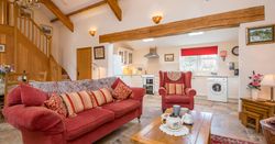 A cozy open-plan living space with a red sofa, armchair, and wooden coffee table set for tea. The room features exposed beams, a staircase, and a bright kitchen in the background.