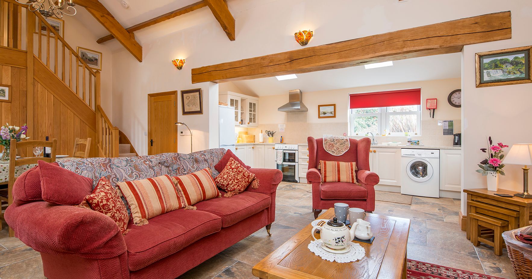 A cozy open-plan living space with a red sofa, armchair, and wooden coffee table set for tea. The room features exposed beams, a staircase, and a bright kitchen in the background.