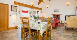 A cozy open-plan dining and living area with wooden beams, a set table with a floral tablecloth, a wood-burning stove, and comfortable seating, leading into a bright kitchen.
