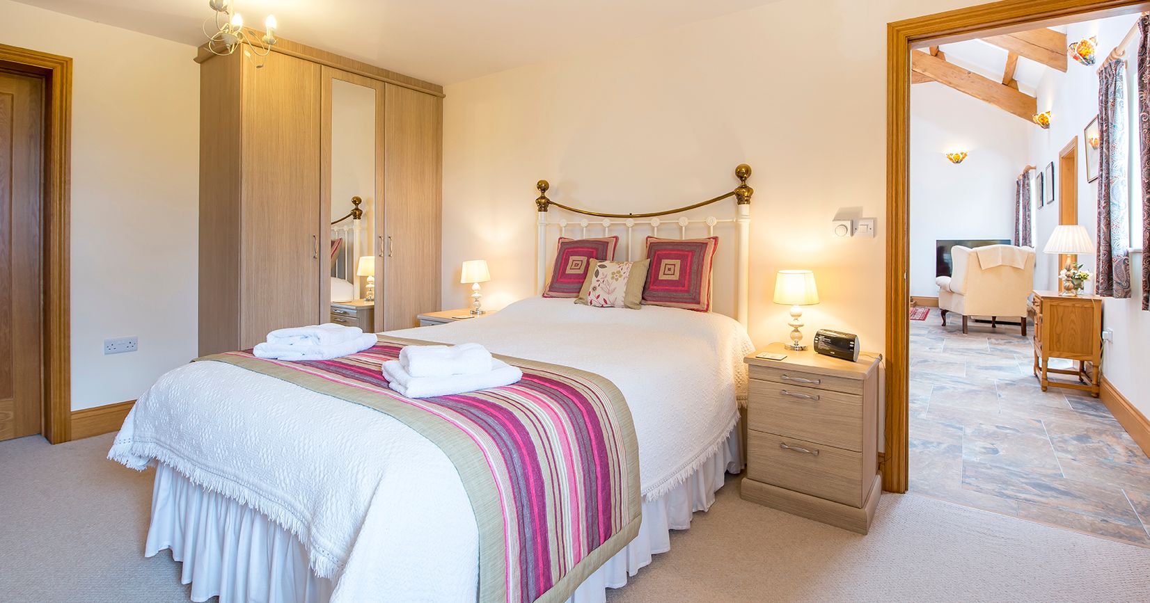 A cozy bedroom with a brass bed frame, white bedding, and a striped throw in shades of pink and green. A wardrobe with mirrors stands beside bedside tables with lamps.