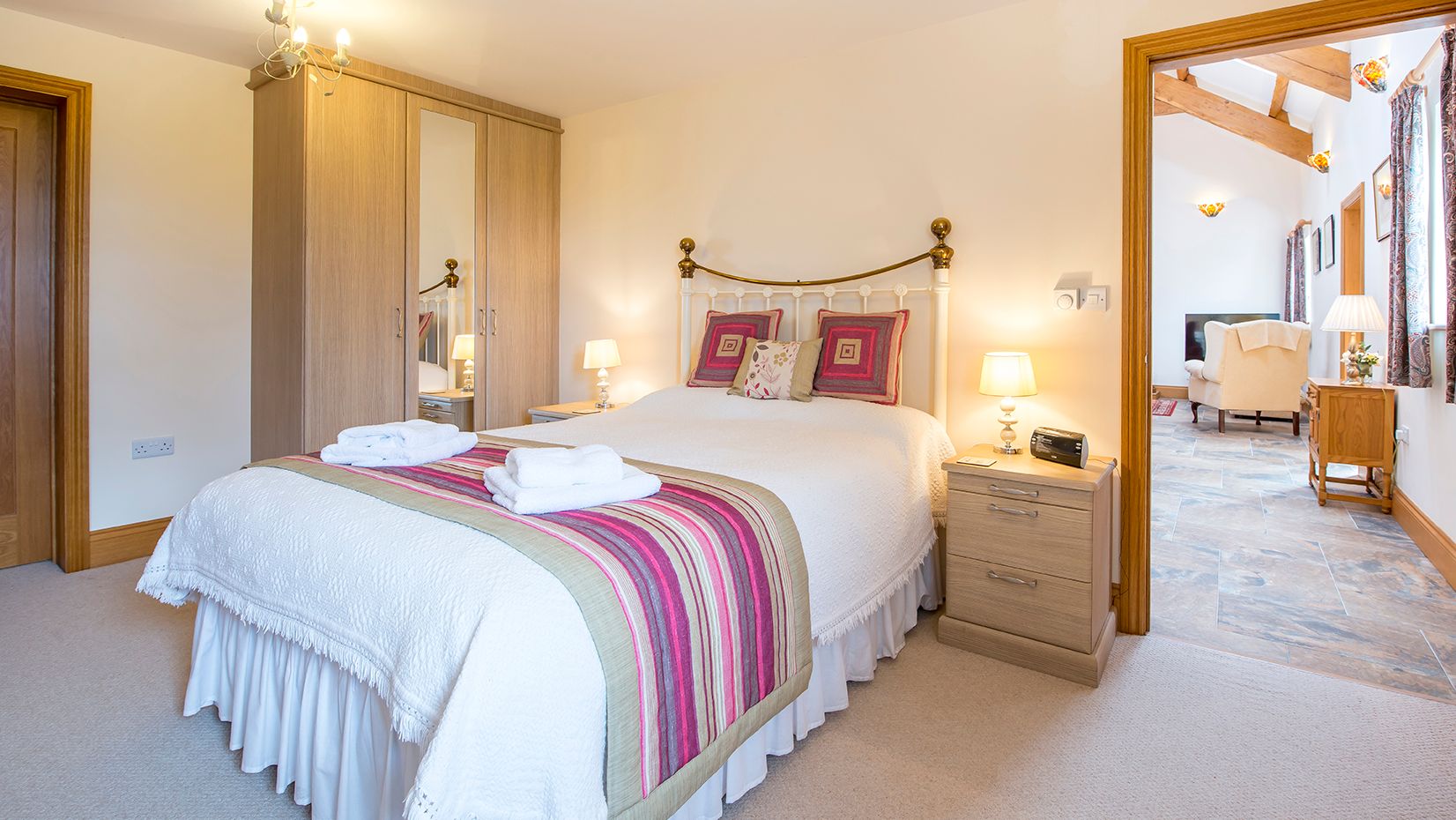A cozy bedroom with a brass bed frame, white bedding, and a striped throw in shades of pink and green. A wardrobe with mirrors stands beside bedside tables with lamps.