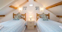 A cozy attic bedroom with two wooden twin beds, dressed in blue and white bedding with matching pillows. A bedside table with a lamp sits between them, under exposed beams.