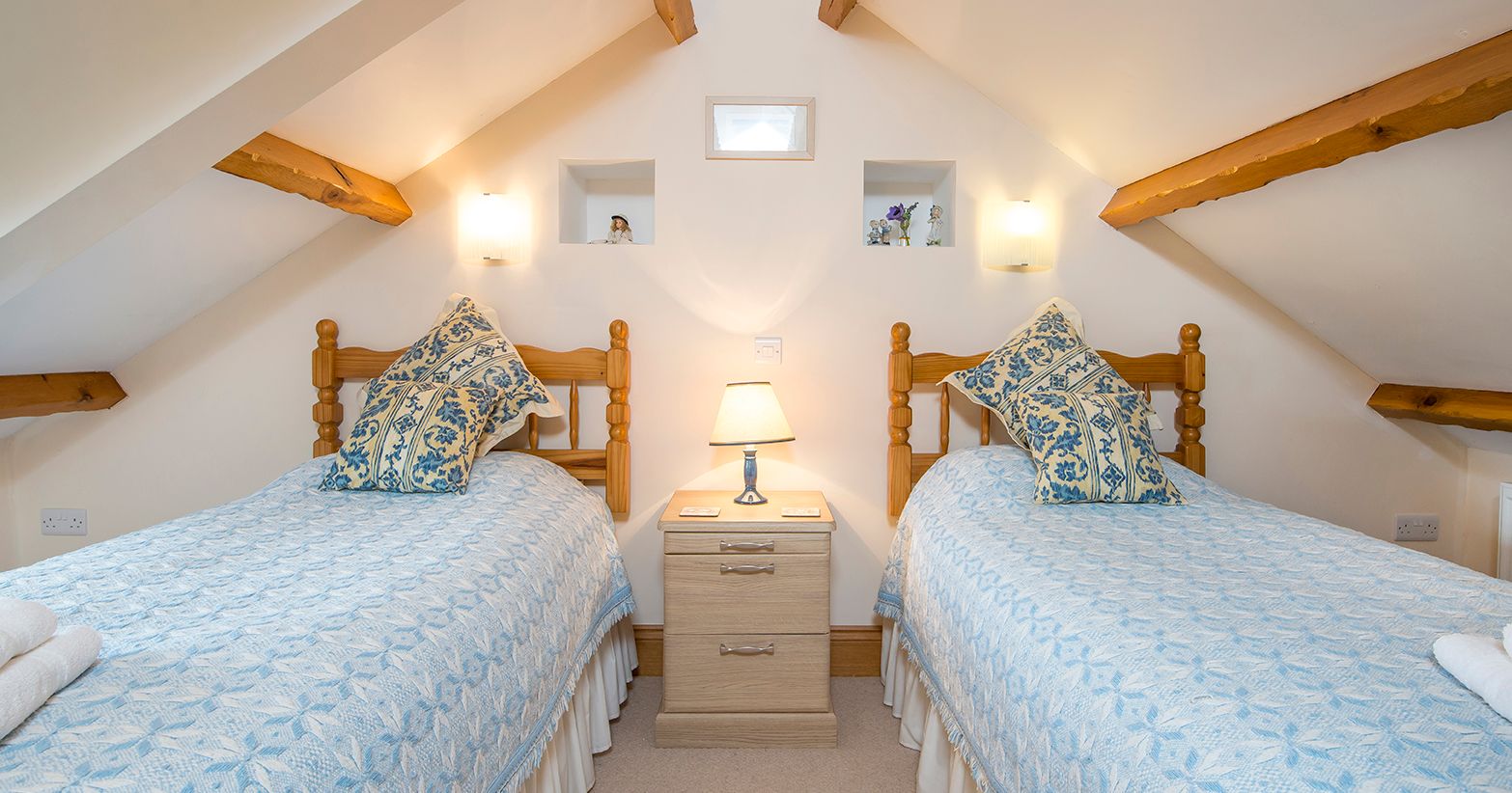A cozy attic bedroom with two wooden twin beds, dressed in blue and white bedding with matching pillows. A bedside table with a lamp sits between them, under exposed beams.