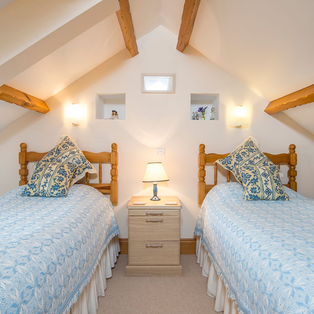 A cozy attic bedroom with two wooden twin beds, dressed in blue and white bedding with matching pillows. A bedside table with a lamp sits between them, under exposed beams.