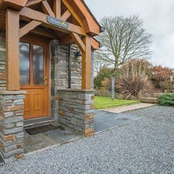 Hirwaun Cottage a rustic stone cottage entrance with a wooden door and porch. A gravel driveway, landscaped garden, and outdoor seating area are nearby.