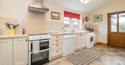 A bright country kitchen with cream cabinets, a white stove, dishwasher, and washing machine. A skylight, window with a red blind, and wooden door add warmth to the space.