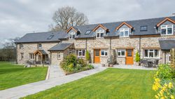 A charming stone-built country house with wooden doors, large windows, and a black slate roof, surrounded by a lush green lawn, vibrant flowers, and outdoor seating areas.