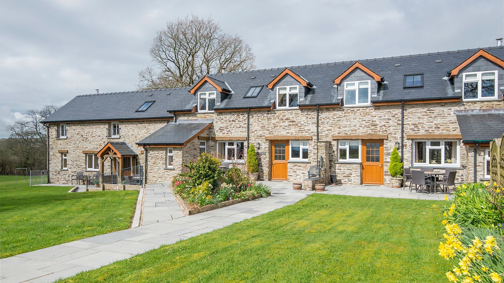 A charming stone-built country house with wooden doors, large windows, and a black slate roof, surrounded by a lush green lawn, vibrant flowers, and outdoor seating areas.