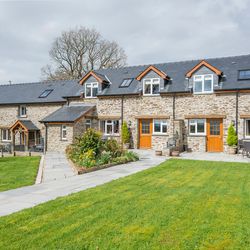A charming stone-built country house with wooden doors, large windows, and a black slate roof, surrounded by a lush green lawn, vibrant flowers, and outdoor seating areas.