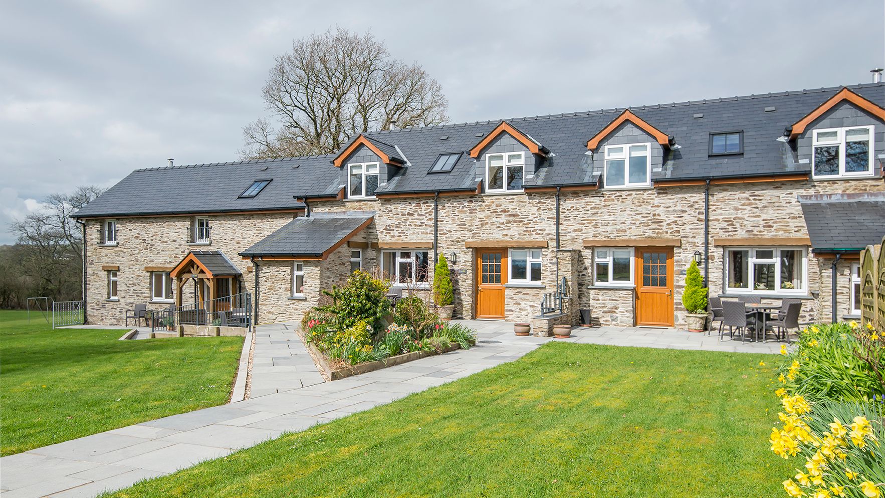 A charming stone-built country house with wooden doors, large windows, and a black slate roof, surrounded by a lush green lawn, vibrant flowers, and outdoor seating areas.