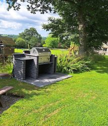 A built-in outdoor barbecue with a stainless steel grill sits on a paved area beside a picnic table, with a wooden playset and a stone house in the lush green background.