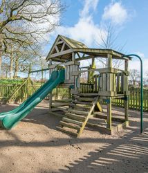 A wooden playground set with a green slide, ladder, and swing frame stands in a sandy, fenced-in area, surrounded by trees and greenery under a bright blue sky.