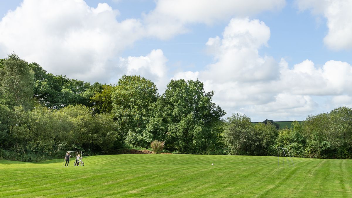 A large, well-manicured grassy field with two small soccer goals, surrounded by lush green trees. A person and a child with a bicycle stand near one of the goals.