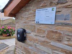 A black electric vehicle charger is mounted on a stone wall beneath a sign with charging instructions. A garden with blooming flowers is visible in the background.