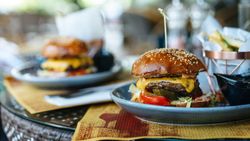 A juicy cheeseburger with lettuce, tomato, pickles, and melted cheese on a sesame seed bun sits on a plate, with fries and another burger in the blurred background.