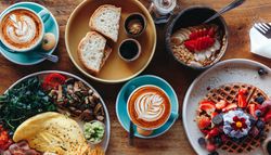 A vibrant breakfast spread featuring lattes, waffles with berries, a smoothie bowl, an omelet with sides, and sourdough toast with butter and jam on a wooden table.