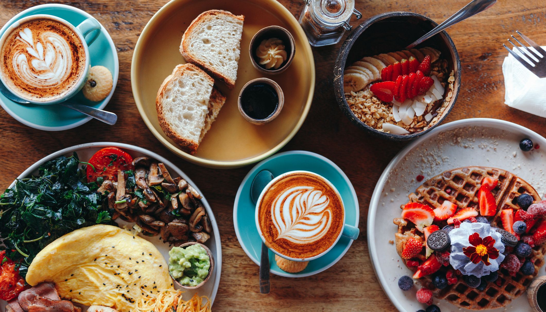 A vibrant breakfast spread featuring lattes, waffles with berries, a smoothie bowl, an omelet with sides, and sourdough toast with butter and jam on a wooden table.