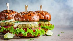 Three cauliflower burgers with seeded brioche buns, lettuce, tomato, cucumber, and creamy sauce are arranged on a rustic surface with lime wedges on the side.