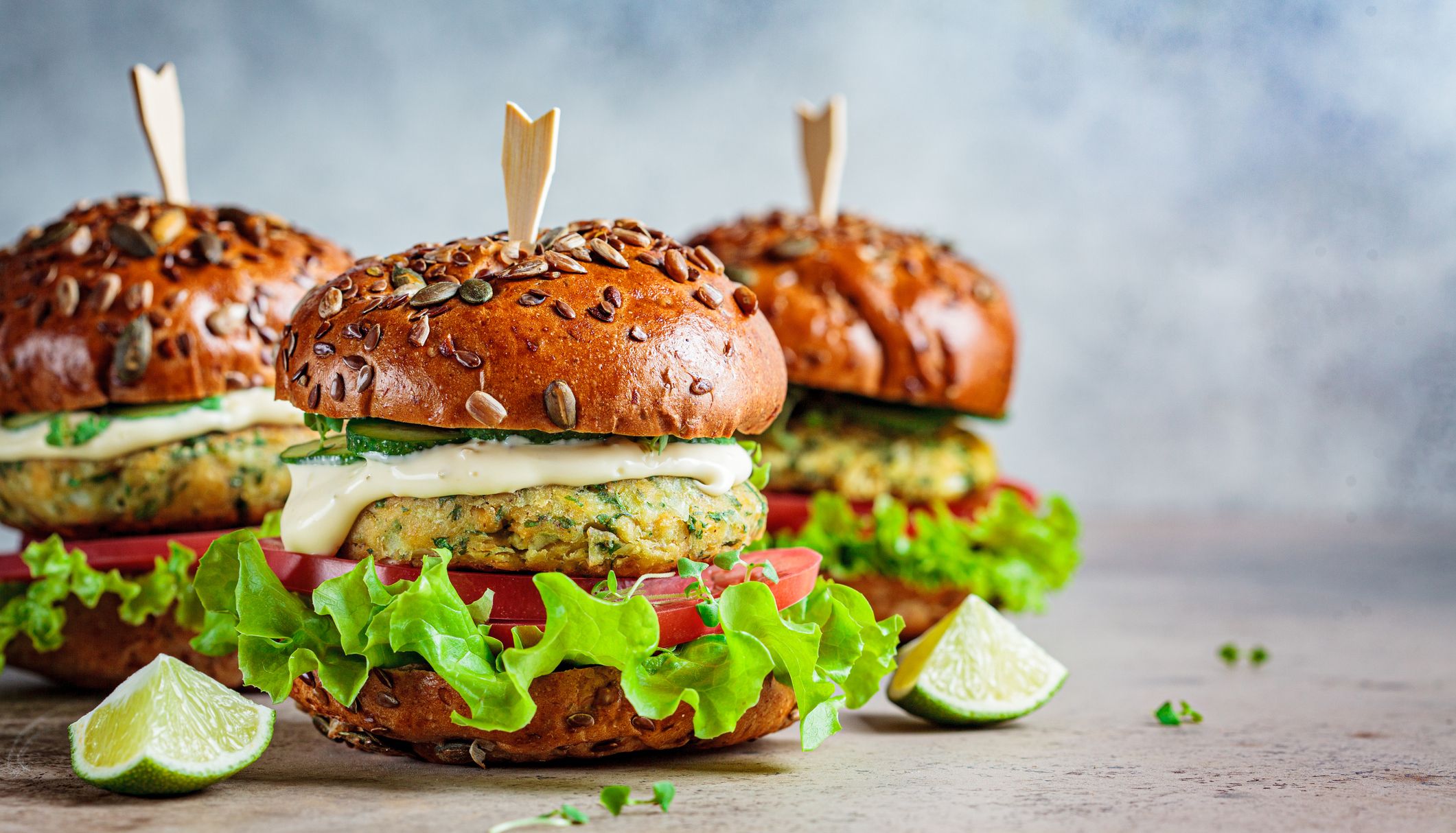 Three cauliflower burgers with seeded brioche buns, lettuce, tomato, cucumber, and creamy sauce are arranged on a rustic surface with lime wedges on the side.