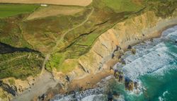 Aerial view of a rugged coastline with steep cliffs, a sandy beach, and turquoise waves crashing against rocky outcrops, with green fields and a small white house above.