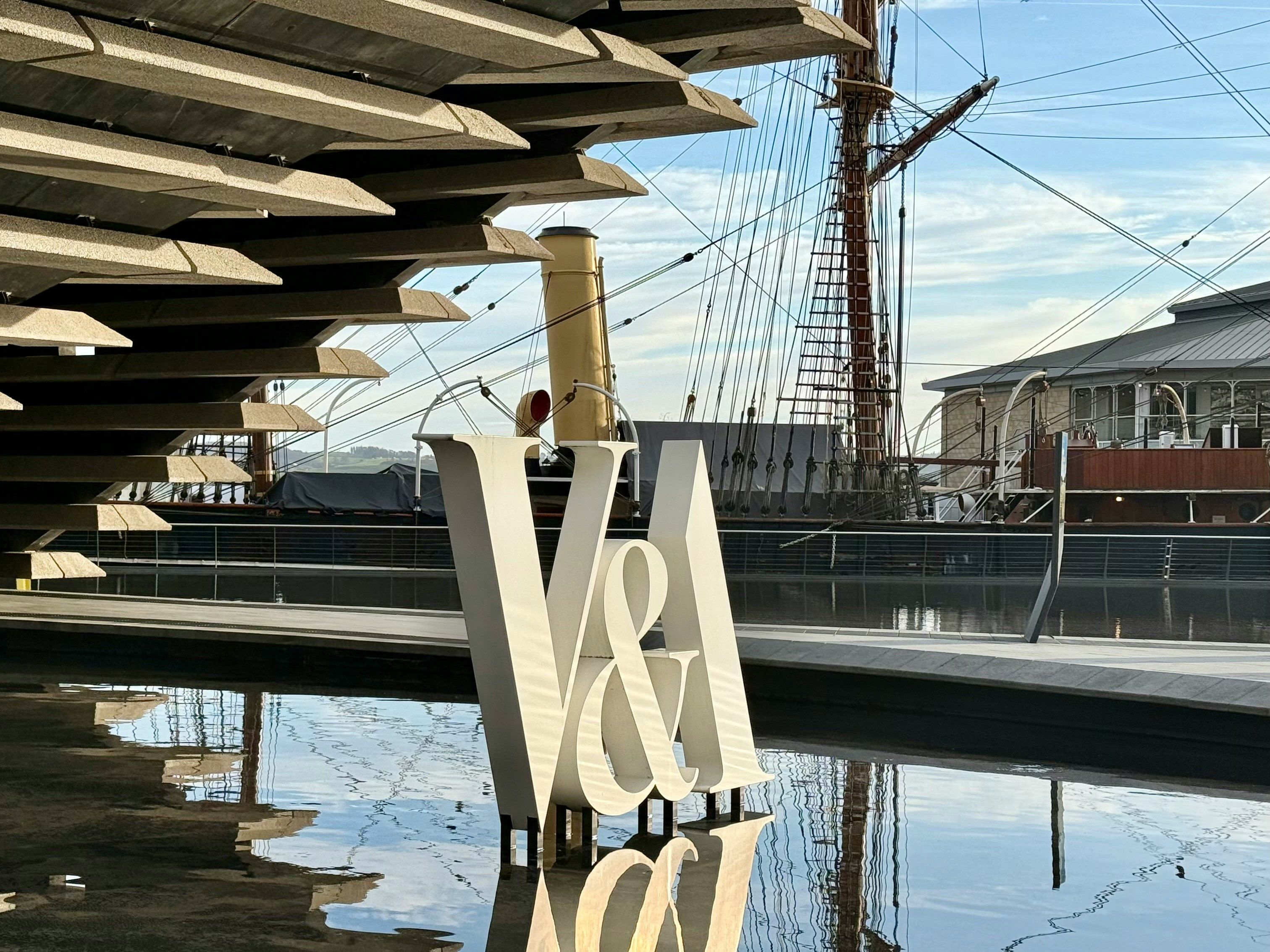 V&A sign reflected in water beside modern terraced building and tall ship rigging