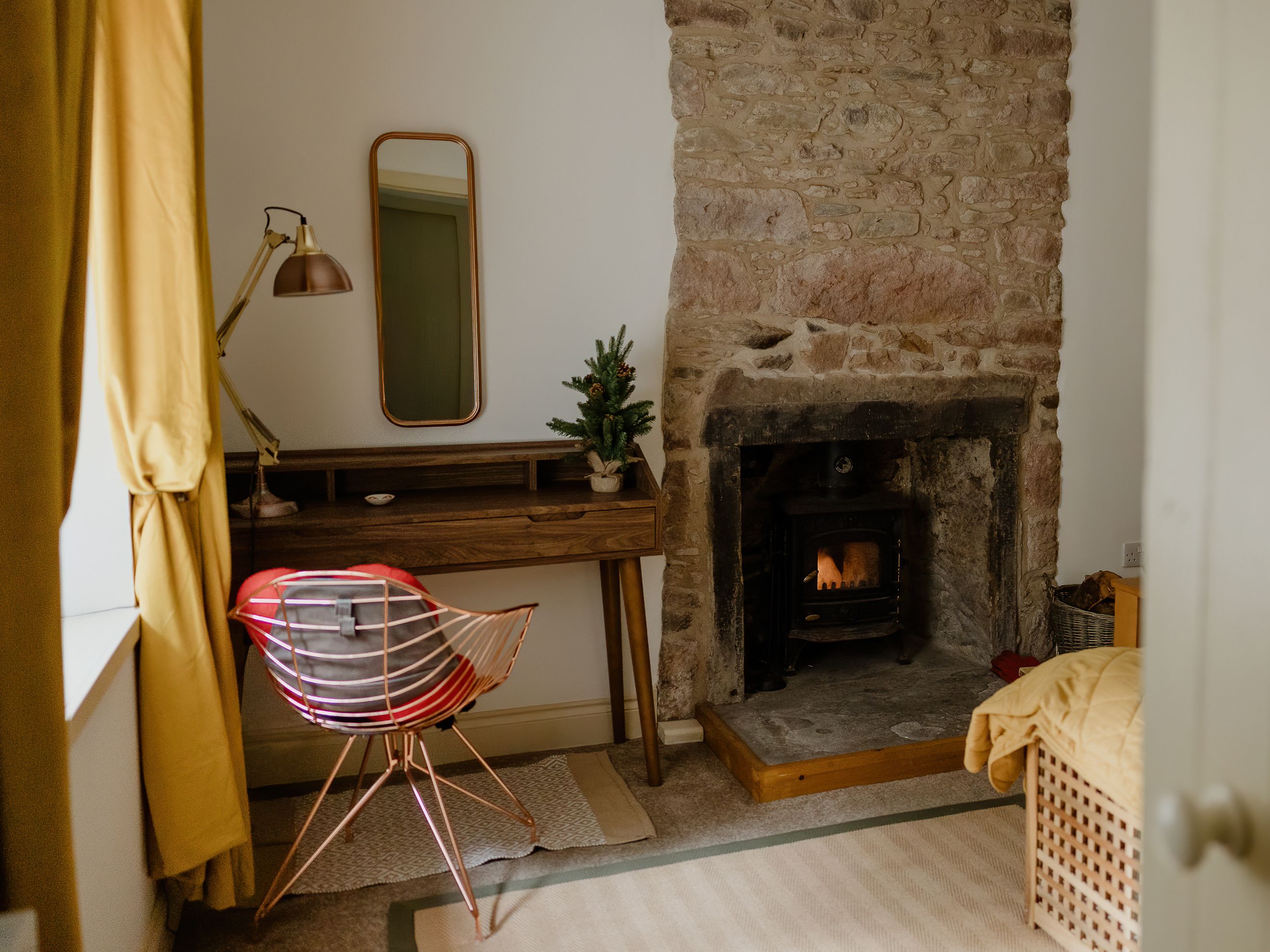 Cosy bedroom corner with stone fireplace, writing desk, mirror and copper chair by a window with mustard curtains