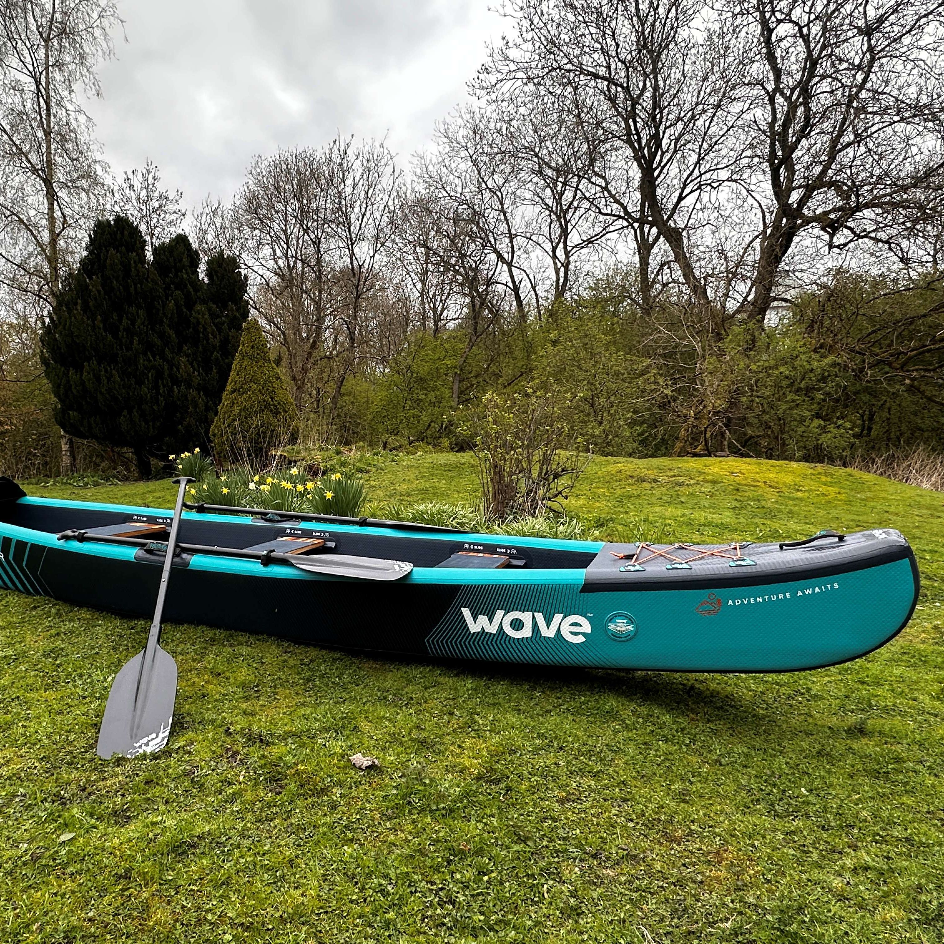 Inflatable turquoise-and-black canoe with paddles on grass in a garden under a cloudy sky