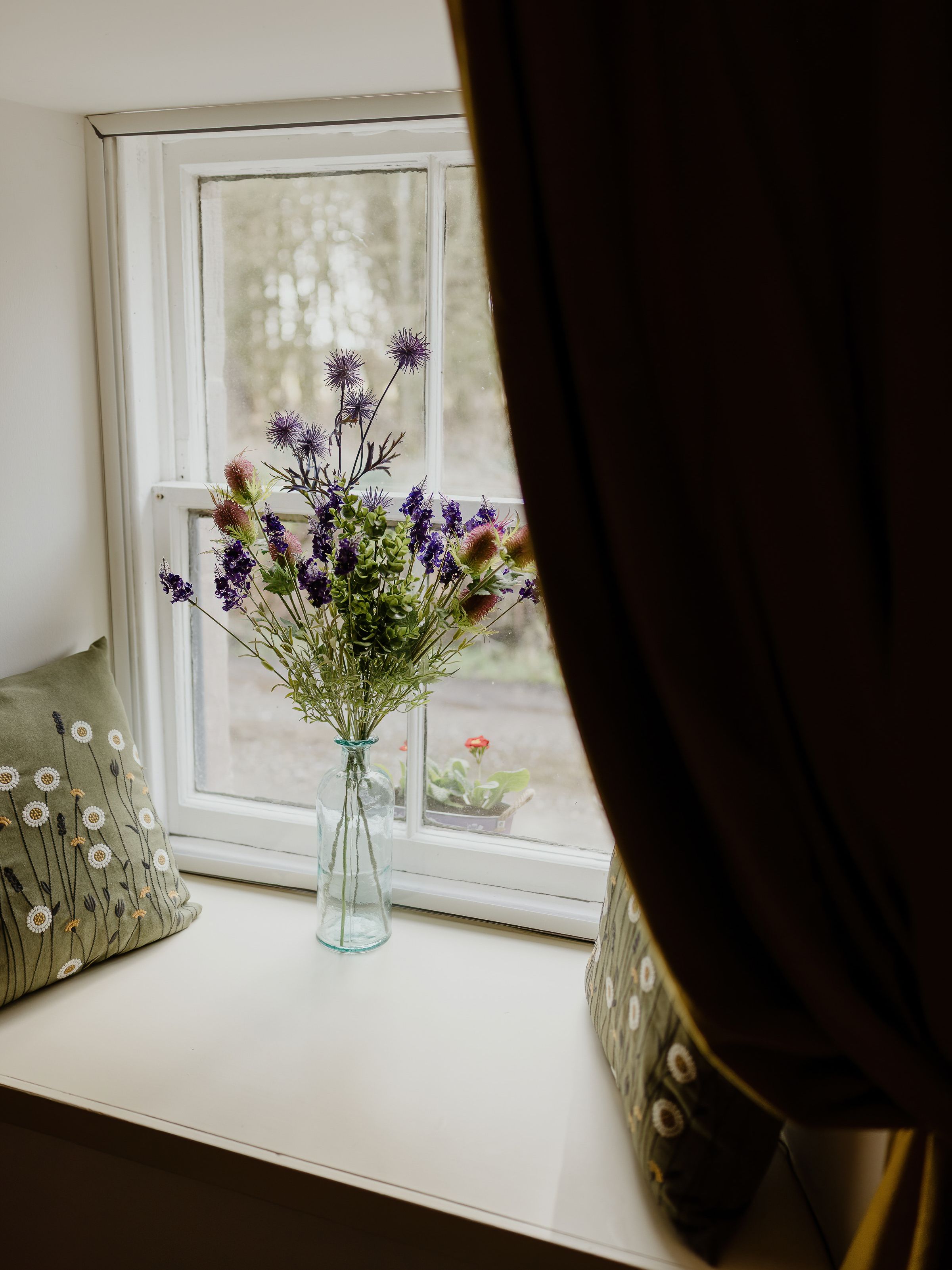 Wildflower bouquet in a glass bottle on a windowsill with patterned cushions and dark curtains