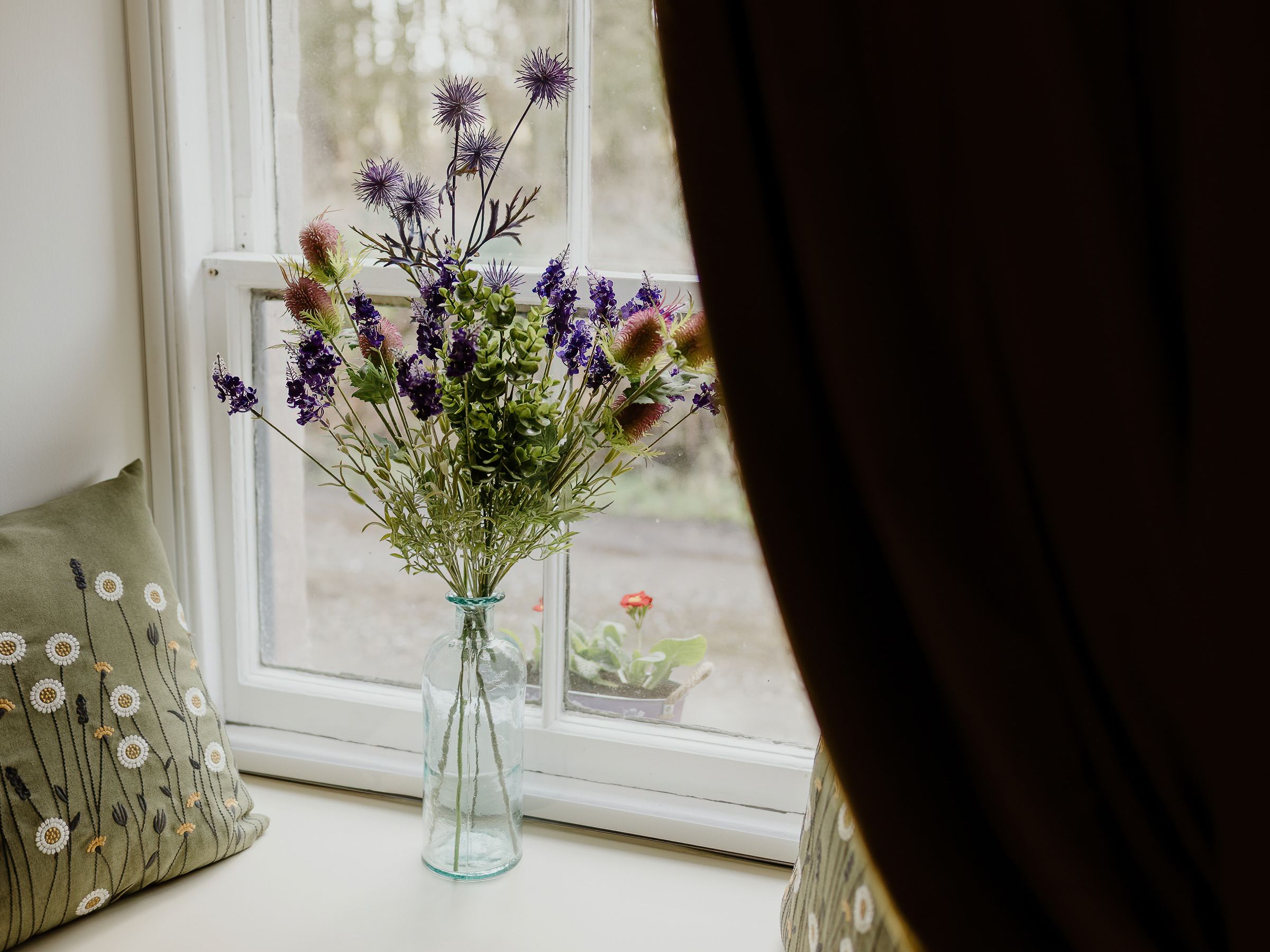 Wildflower bouquet in a glass bottle on a windowsill with patterned cushions and dark curtains