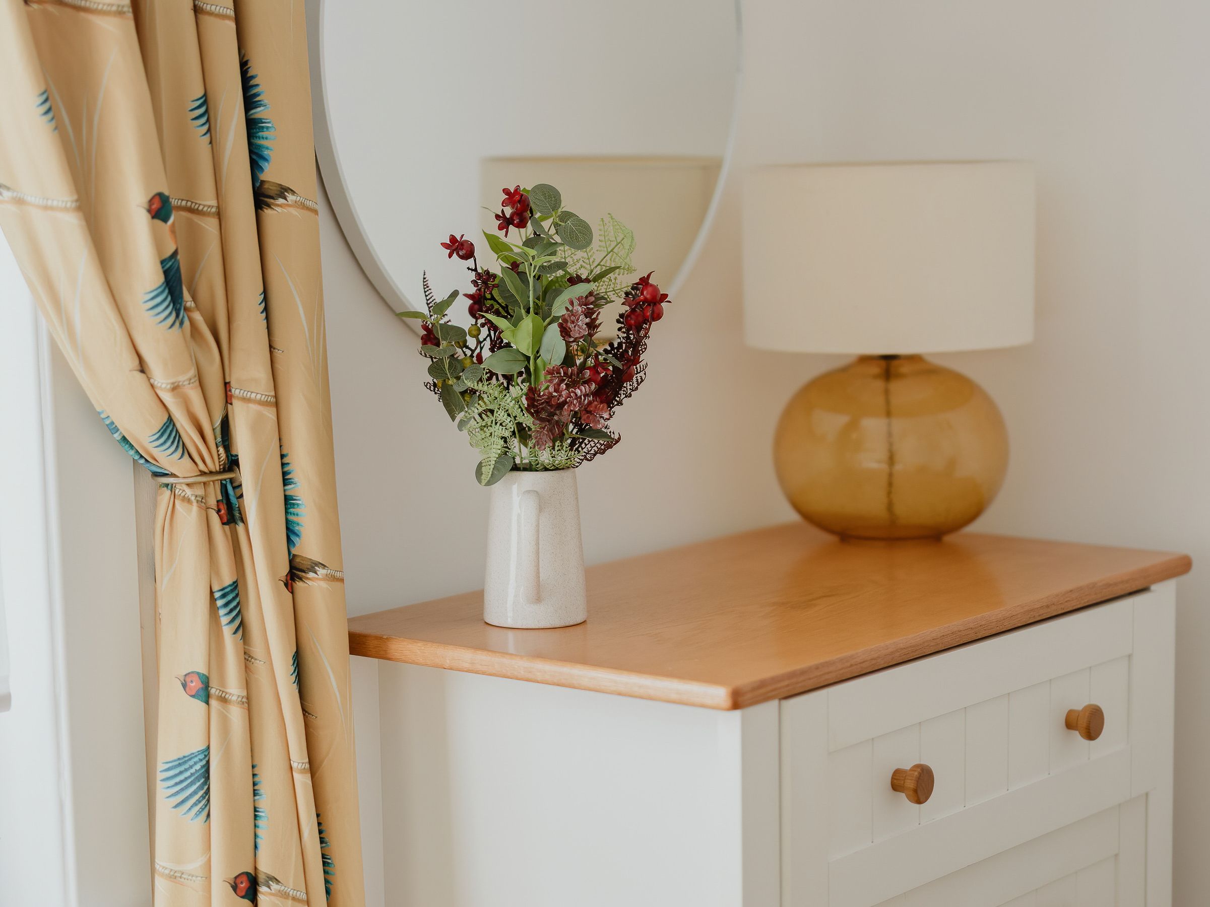 White chest of drawers with a wooden top, round wall mirror, vase of flowers and amber lamp beside patterned curtains