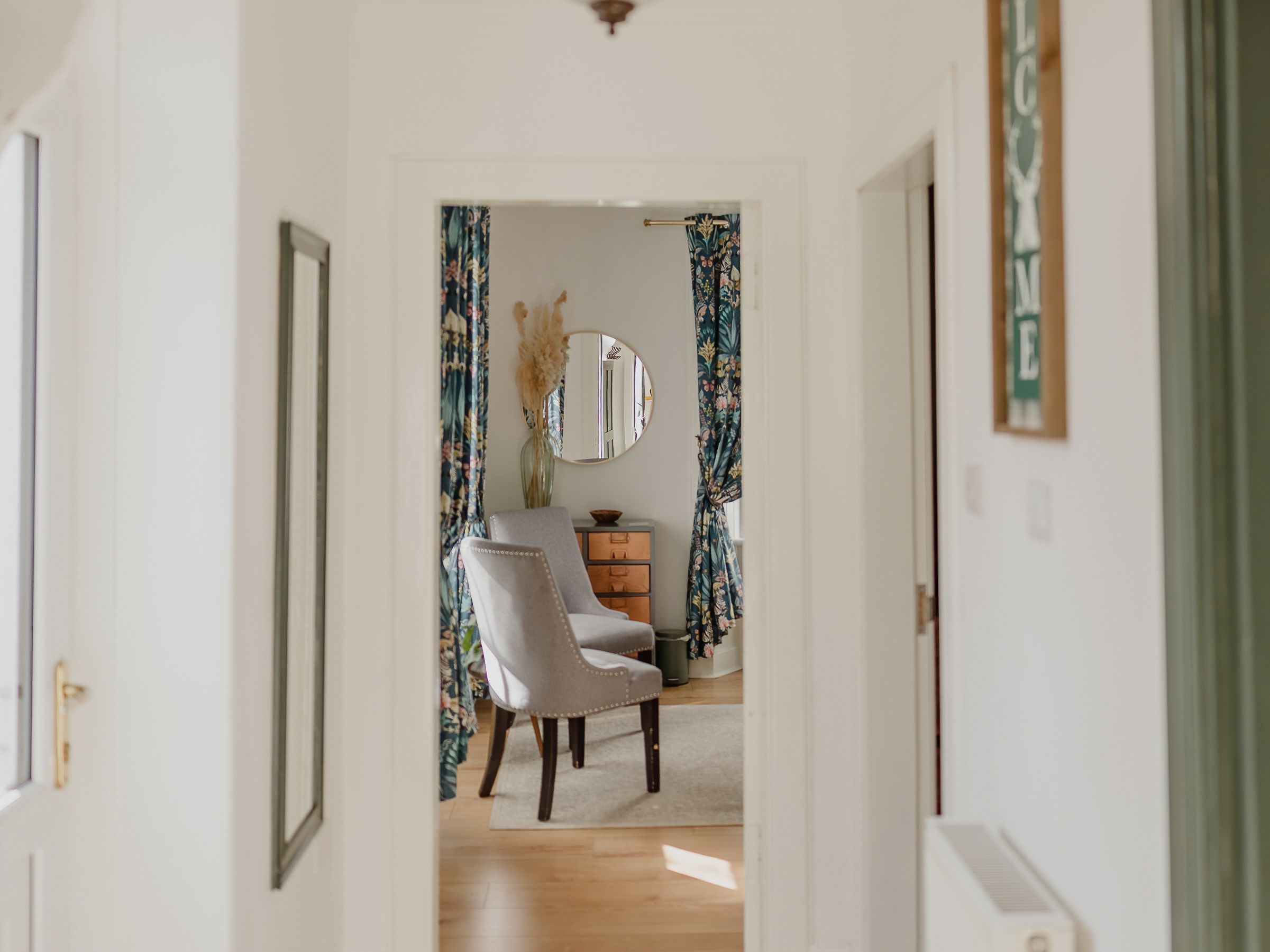 Bright white hallway with hanging pendant light and a doorway to a sitting area with floral curtains and a grey chair