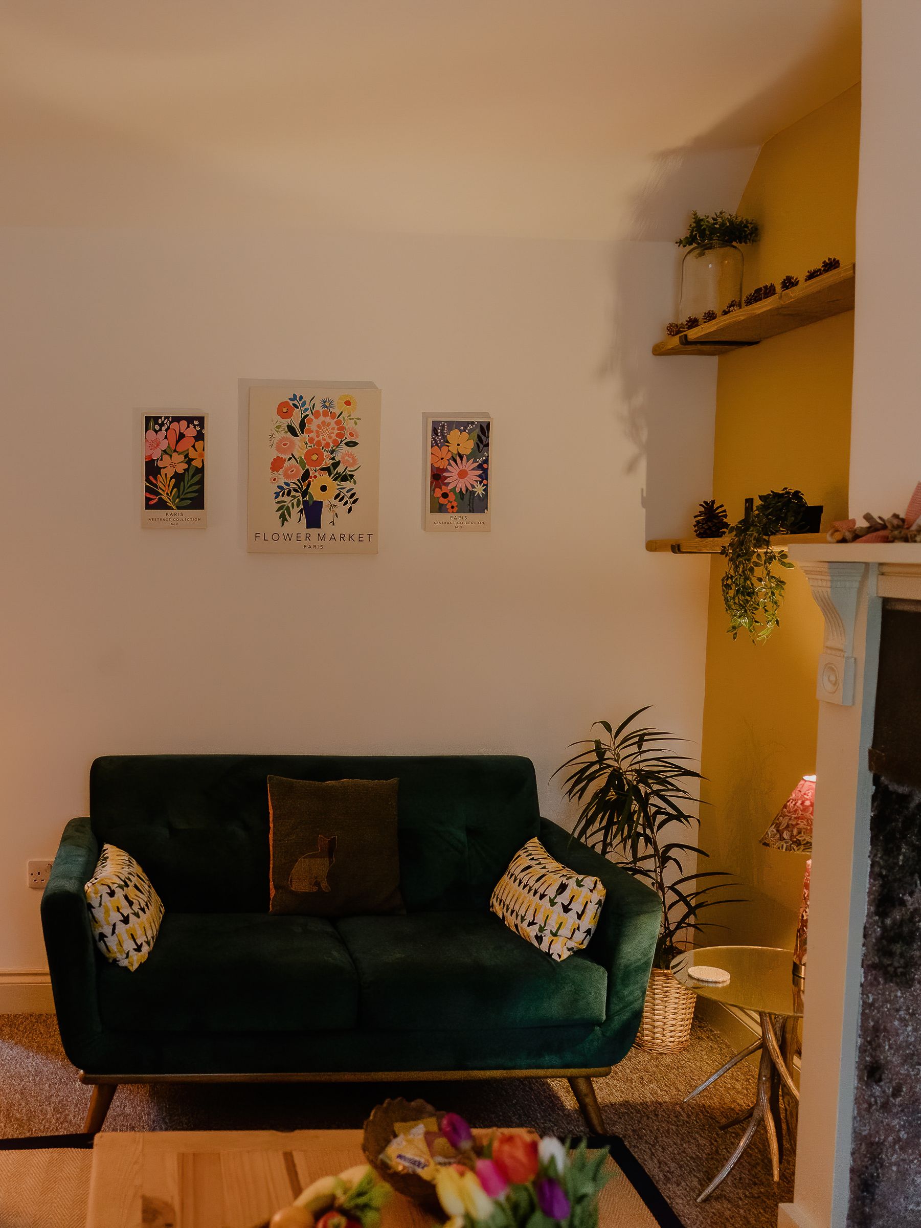 Cosy living room with green velvet sofa, floor lamp, floral wall art, and doorway to a small kitchen beside a fireplace.