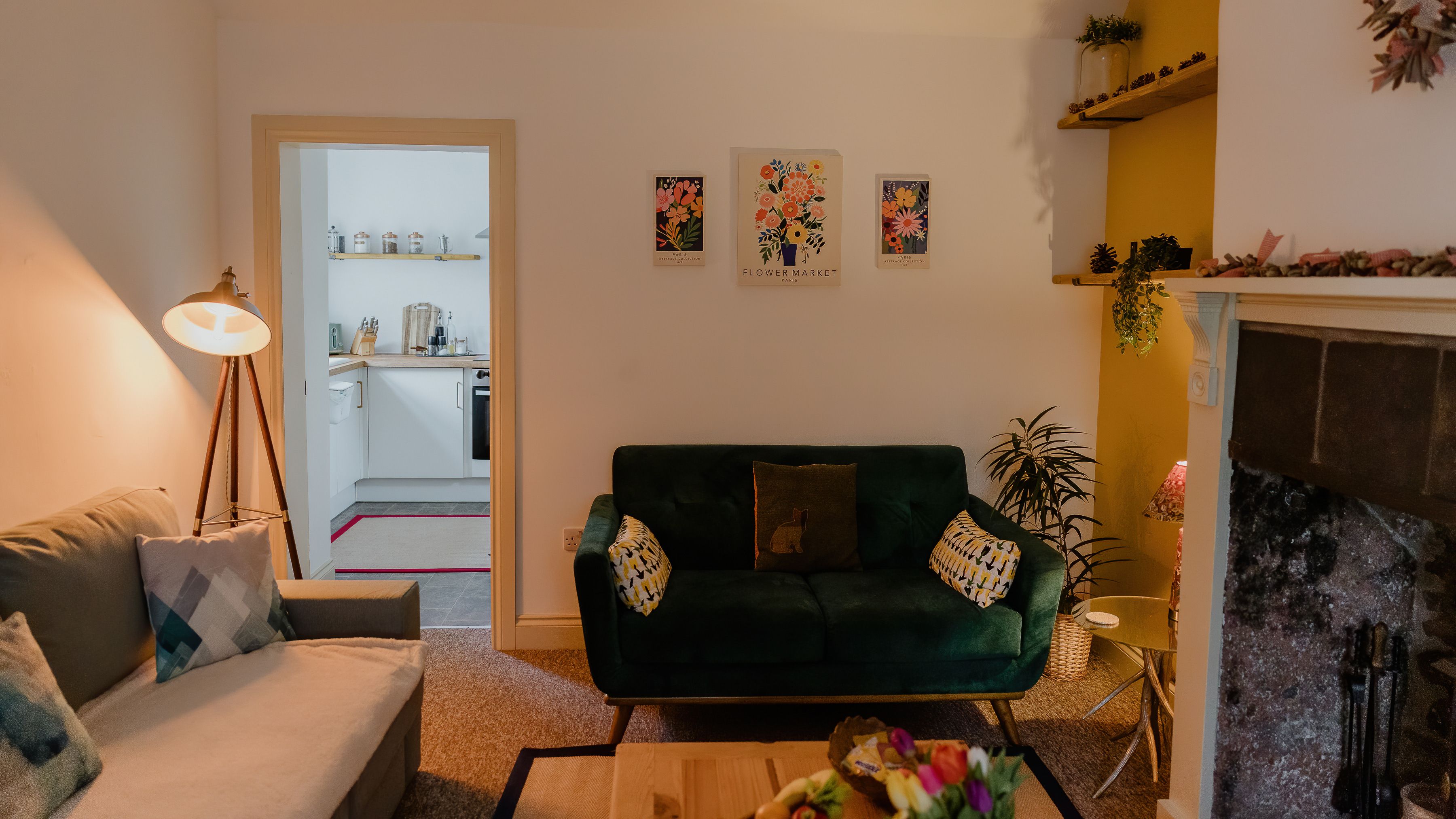 Cosy living room with green velvet sofa, floor lamp, floral wall art, and doorway to a small kitchen beside a fireplace.