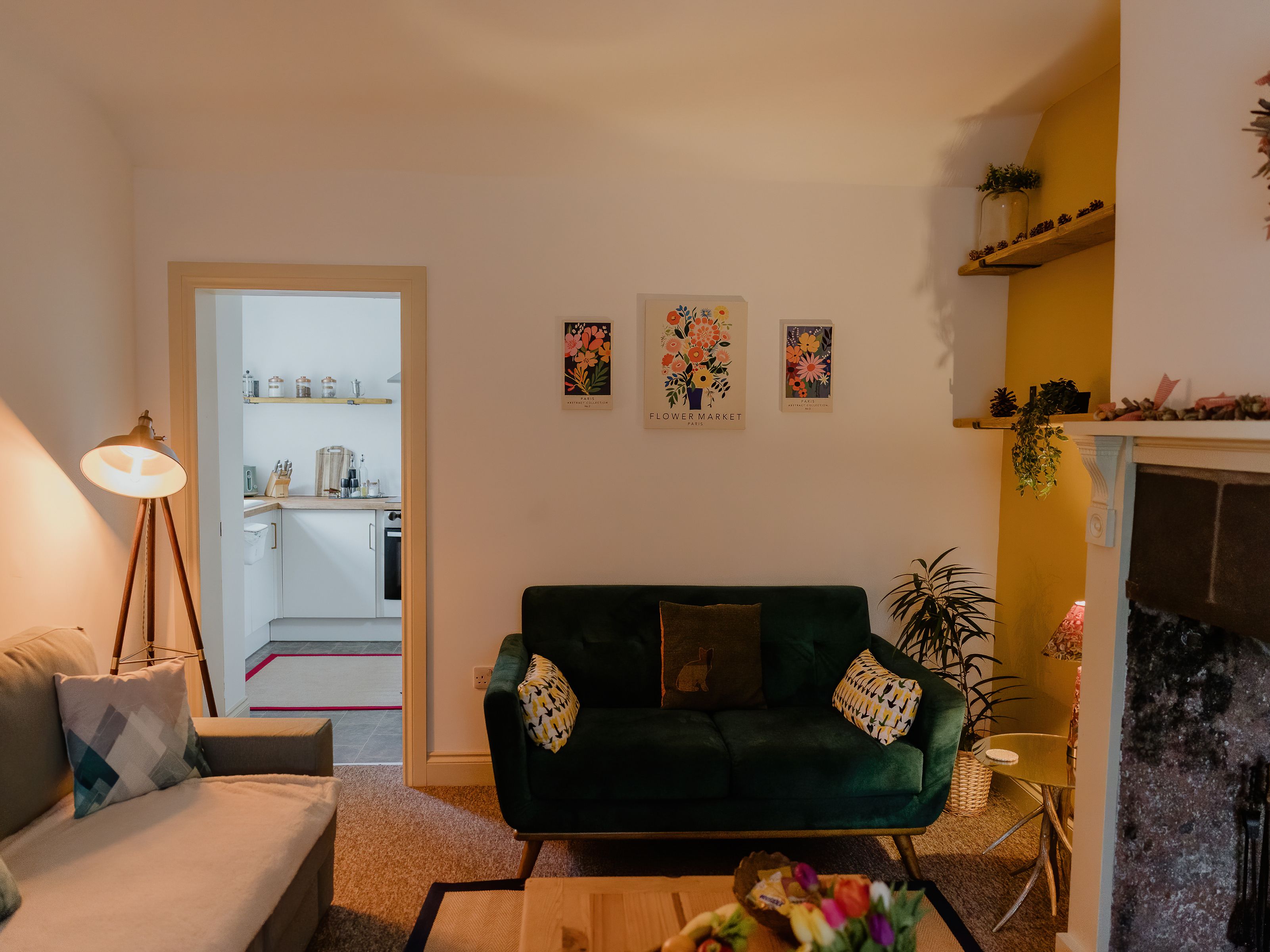 Cosy living room with green velvet sofa, floor lamp, floral wall art, and doorway to a small kitchen beside a fireplace.
