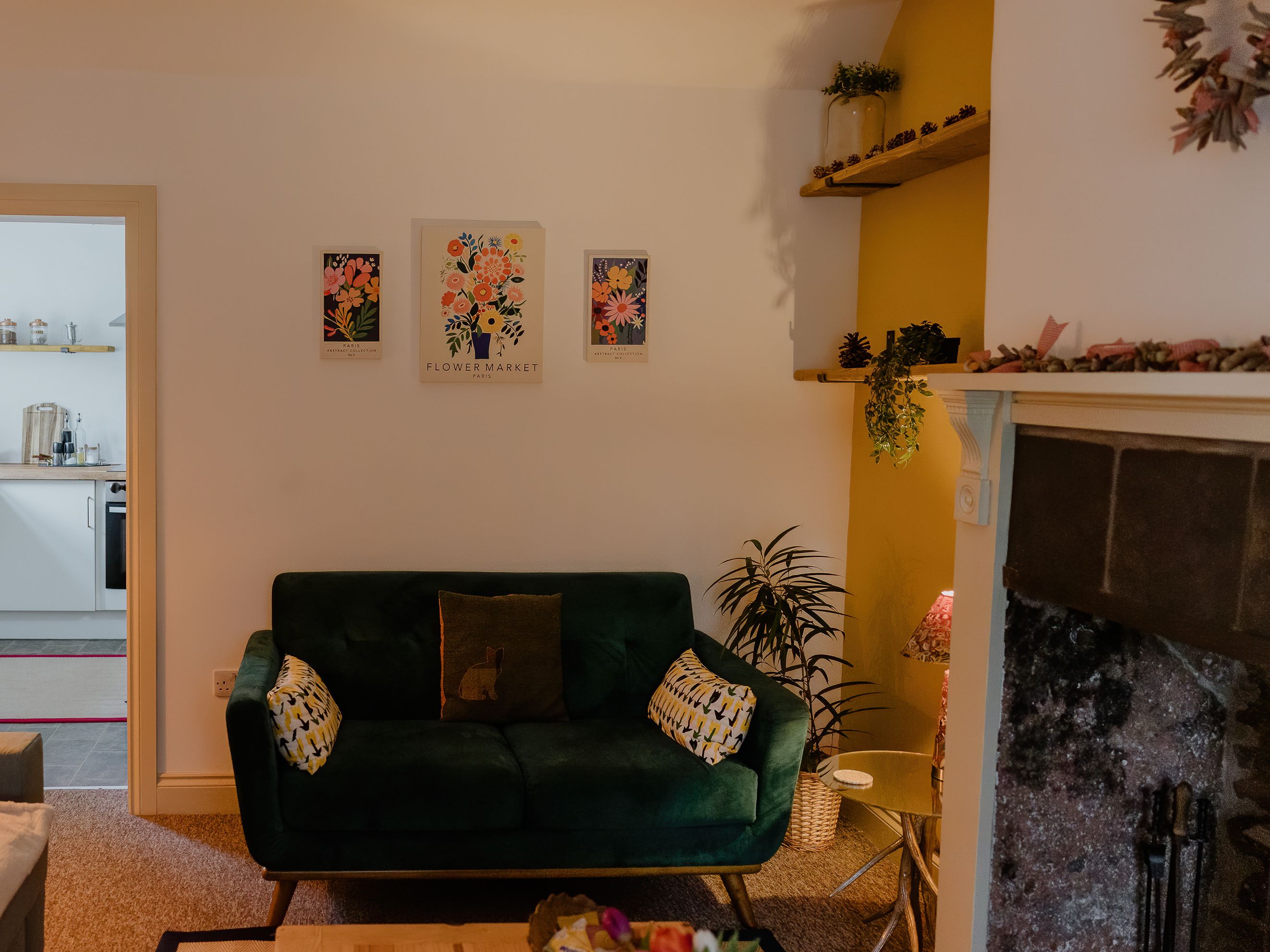 Cosy living room with green velvet sofa, floor lamp, floral wall art, and doorway to a small kitchen beside a fireplace.
