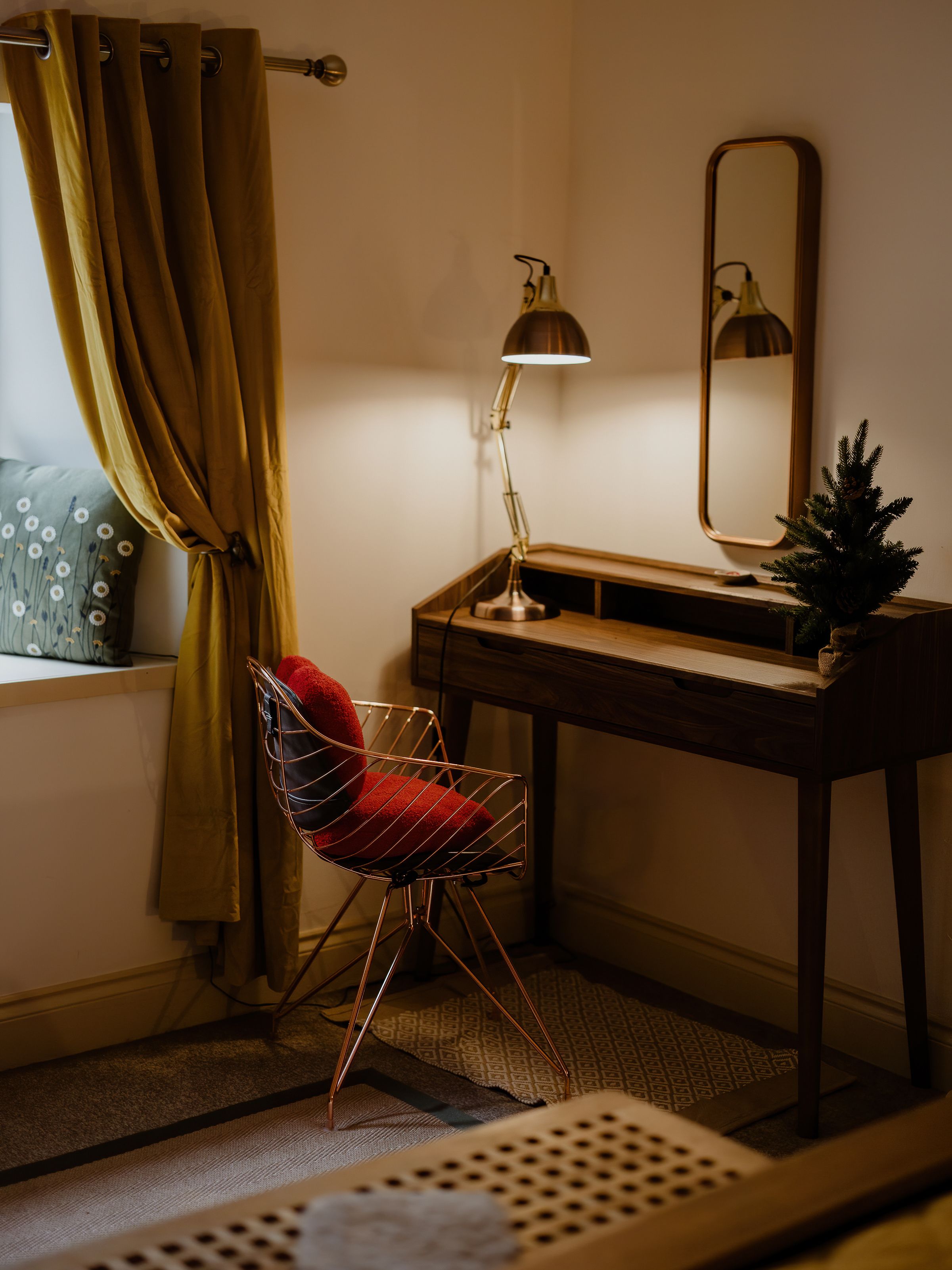Cosy corner with wooden desk, lit task lamp, mirror and wire chair beside mustard curtains