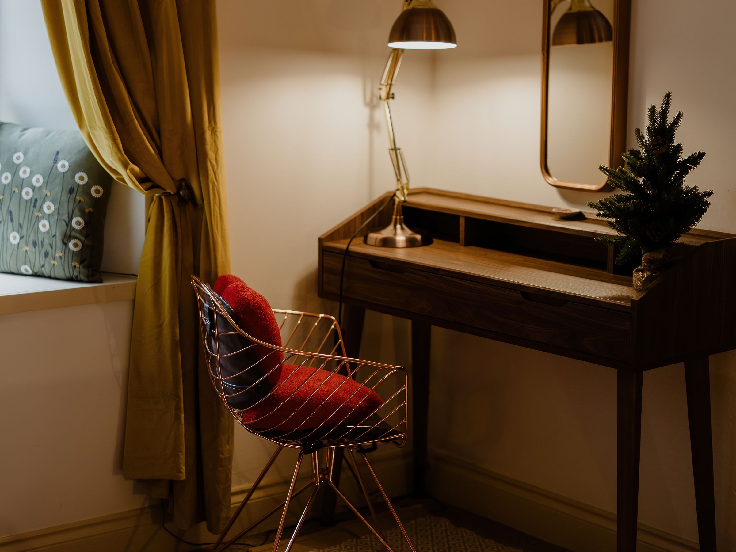 Cosy corner with wooden desk, lit task lamp, mirror and wire chair beside mustard curtains
