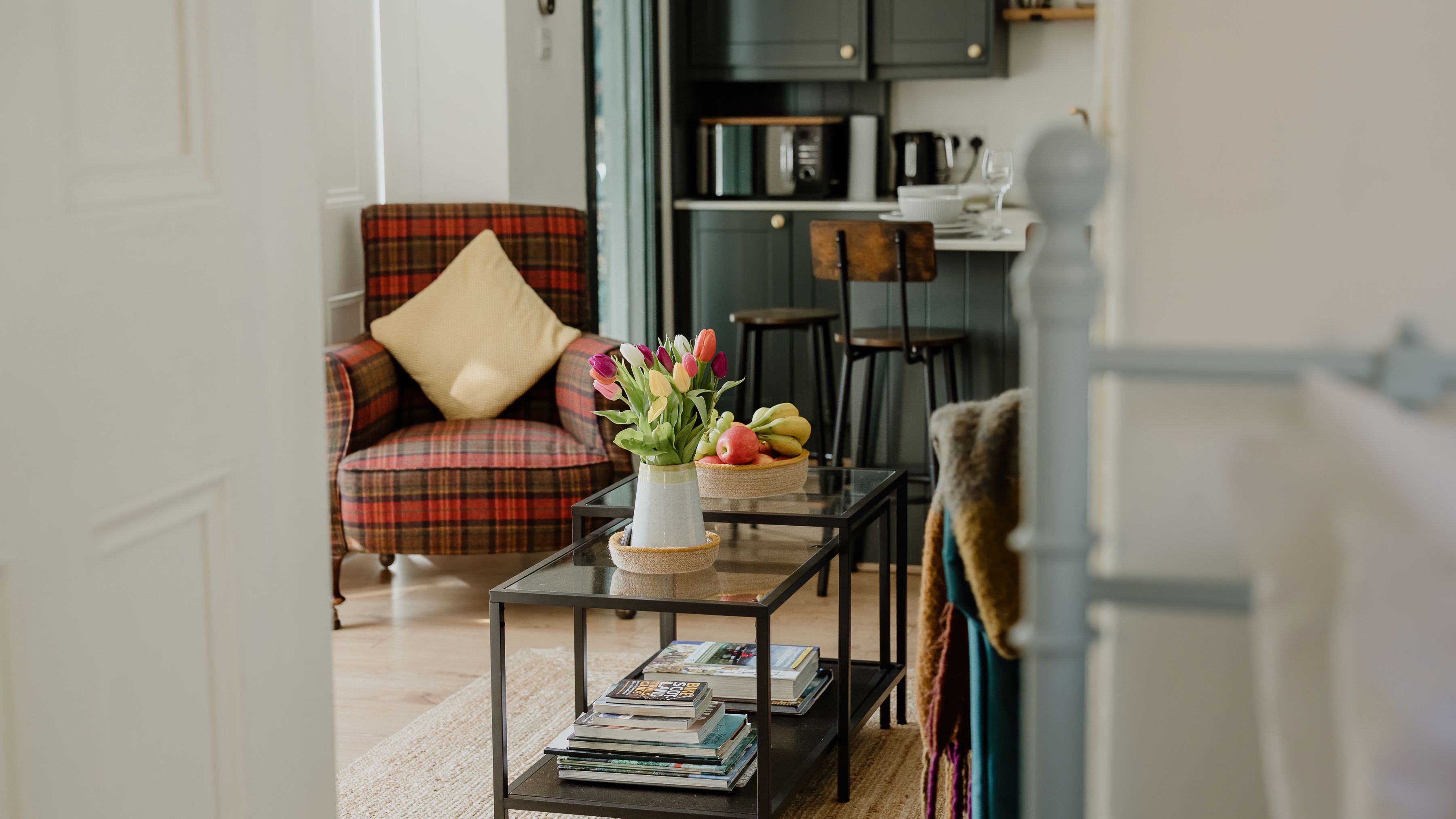 Cosy living room with a tartan armchair, glass coffee tables and tulips, opening onto a small kitchen.