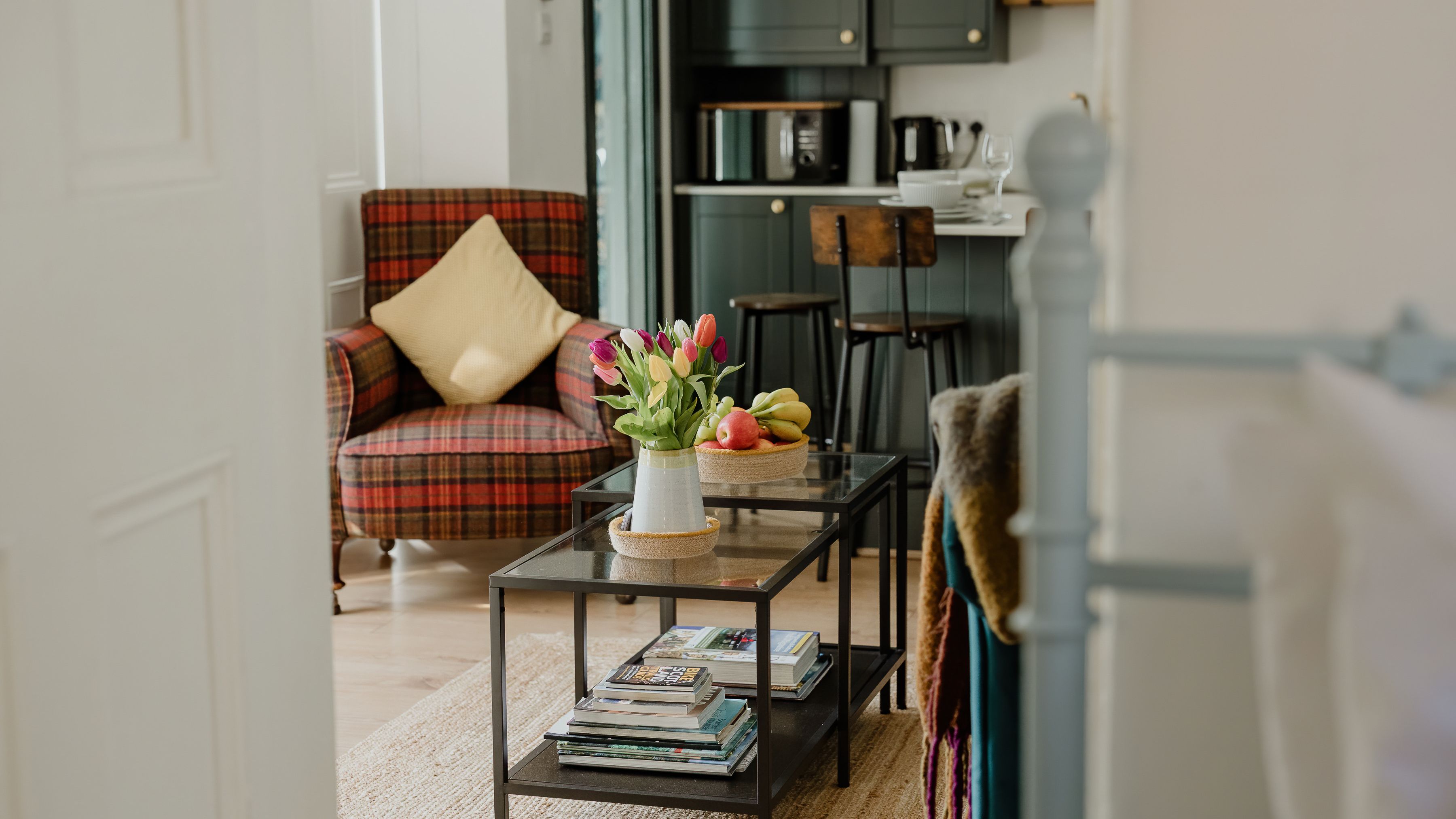 Cosy living room with a tartan armchair, glass coffee tables and tulips, opening onto a small kitchen.