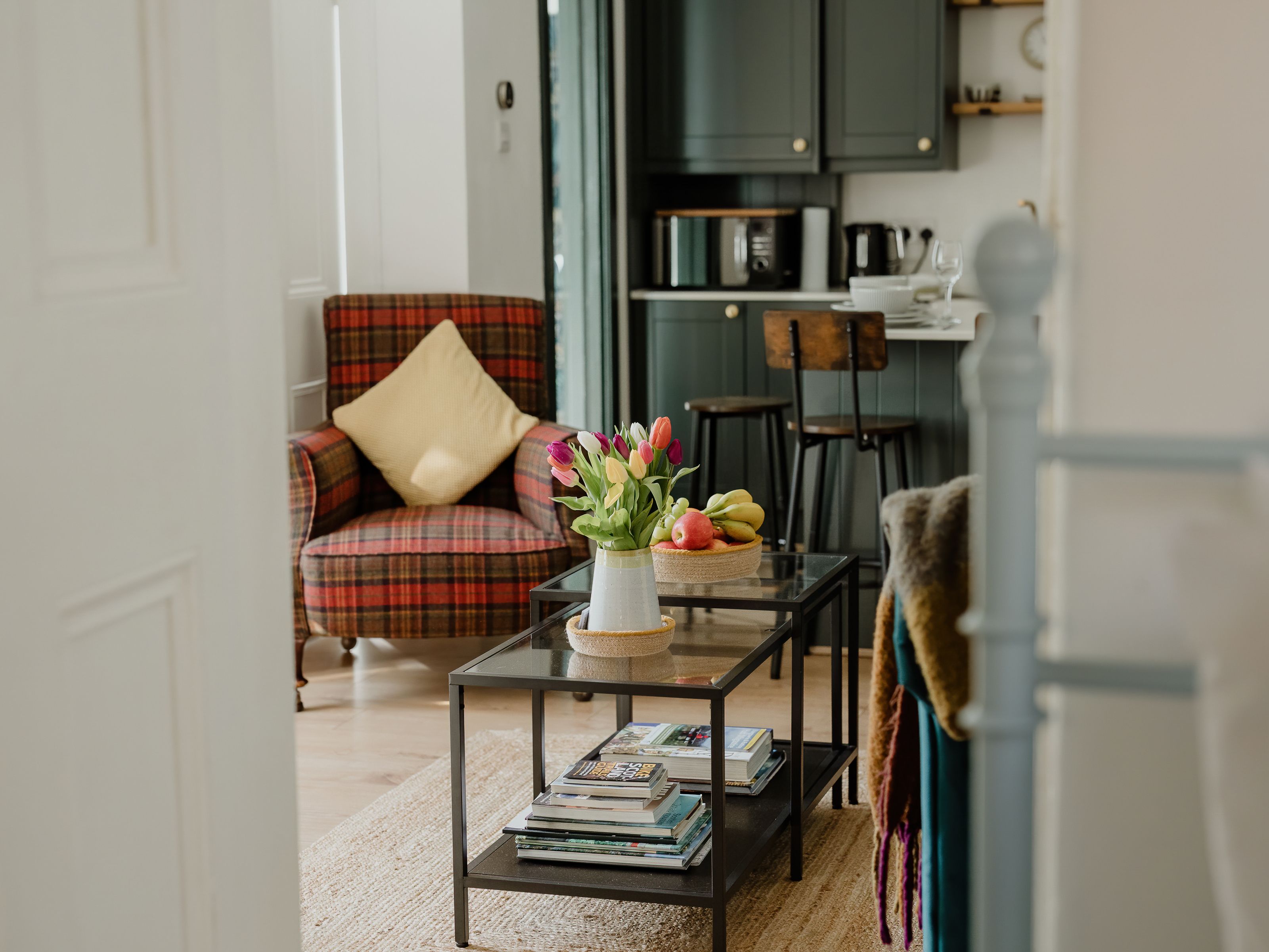Cosy living room with a tartan armchair, glass coffee tables and tulips, opening onto a small kitchen.