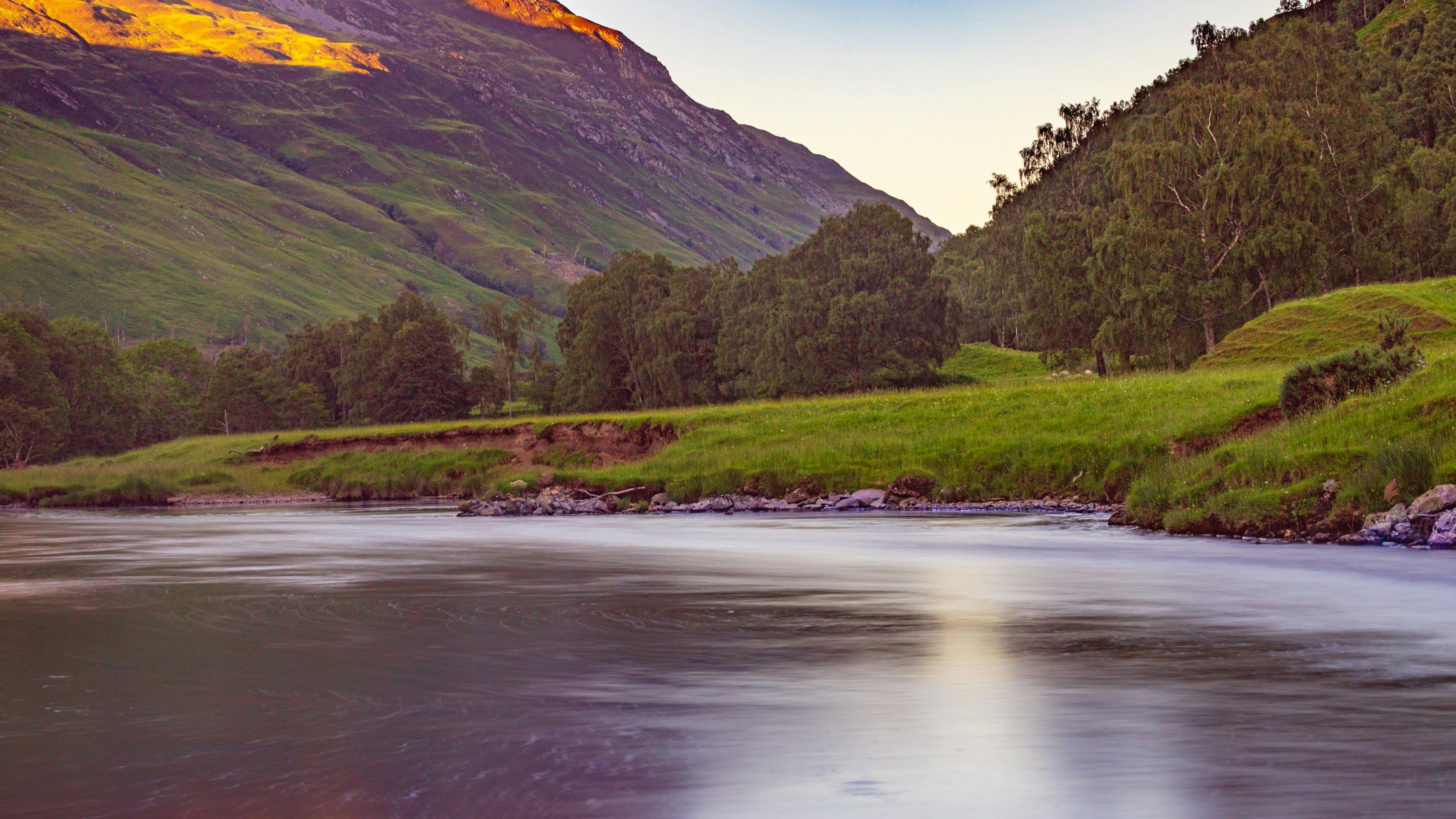 Sunlit mountain slope above a calm river with green meadow and trees