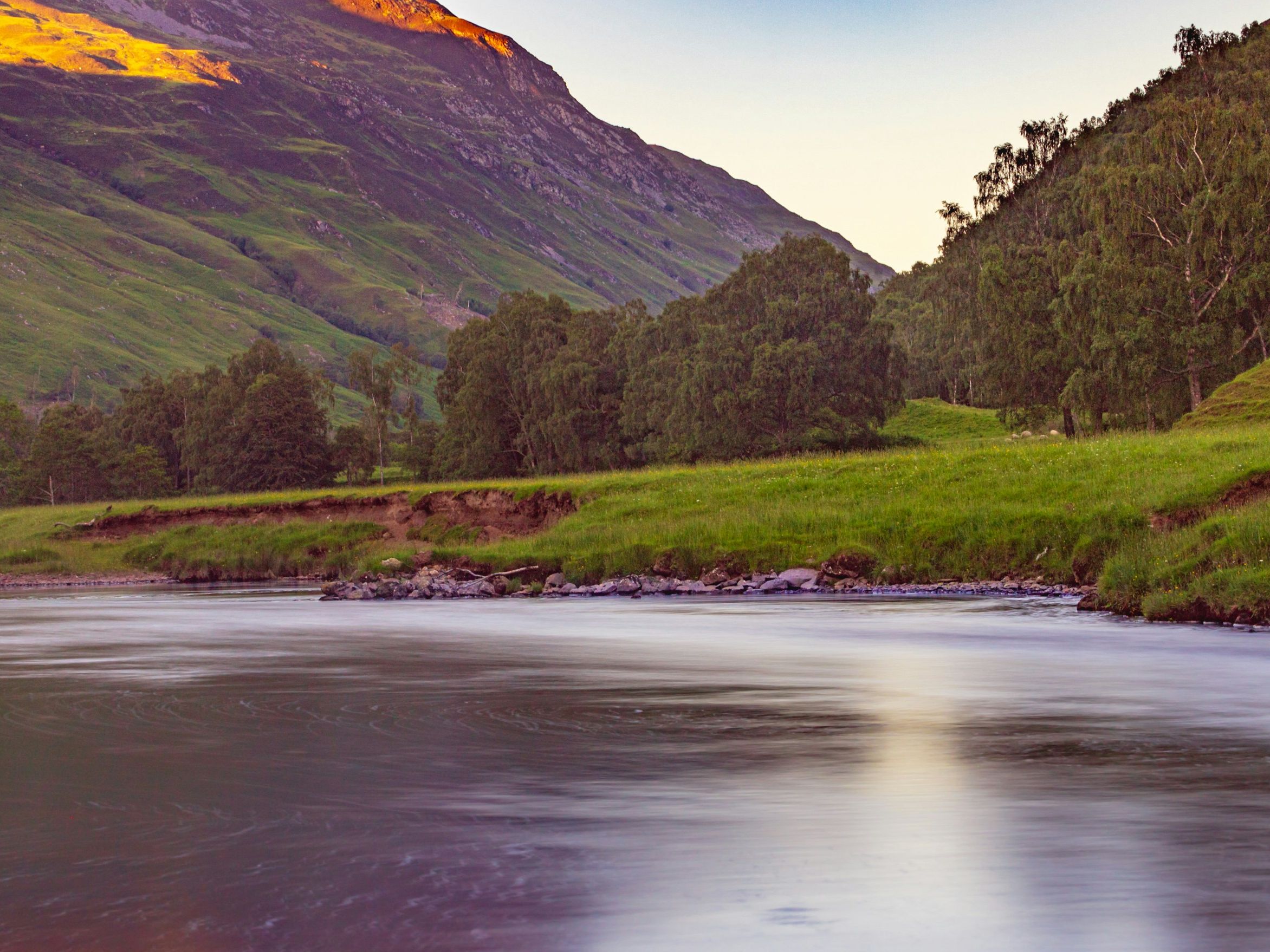 Sunlit mountain slope above a calm river with green meadow and trees