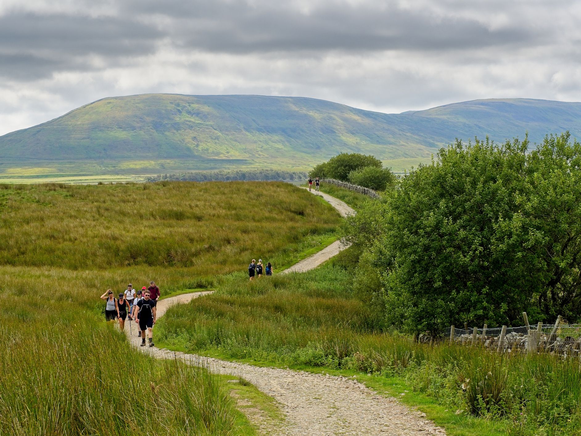 Walkers on a winding path through green countryside with hills under cloudy skies