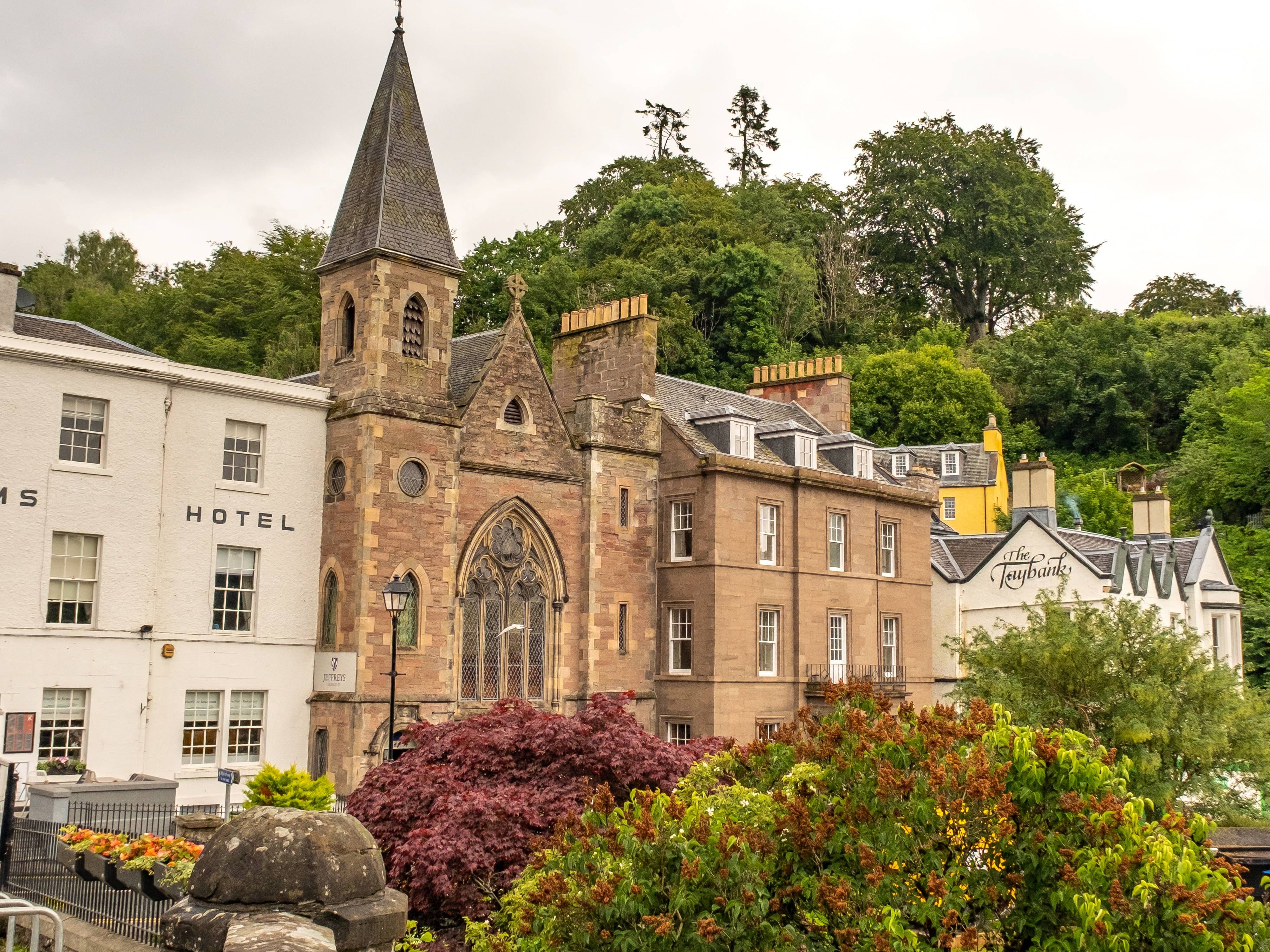 Stone church-like building with a tall spire beside a hotel, viewed over a stone wall and garden