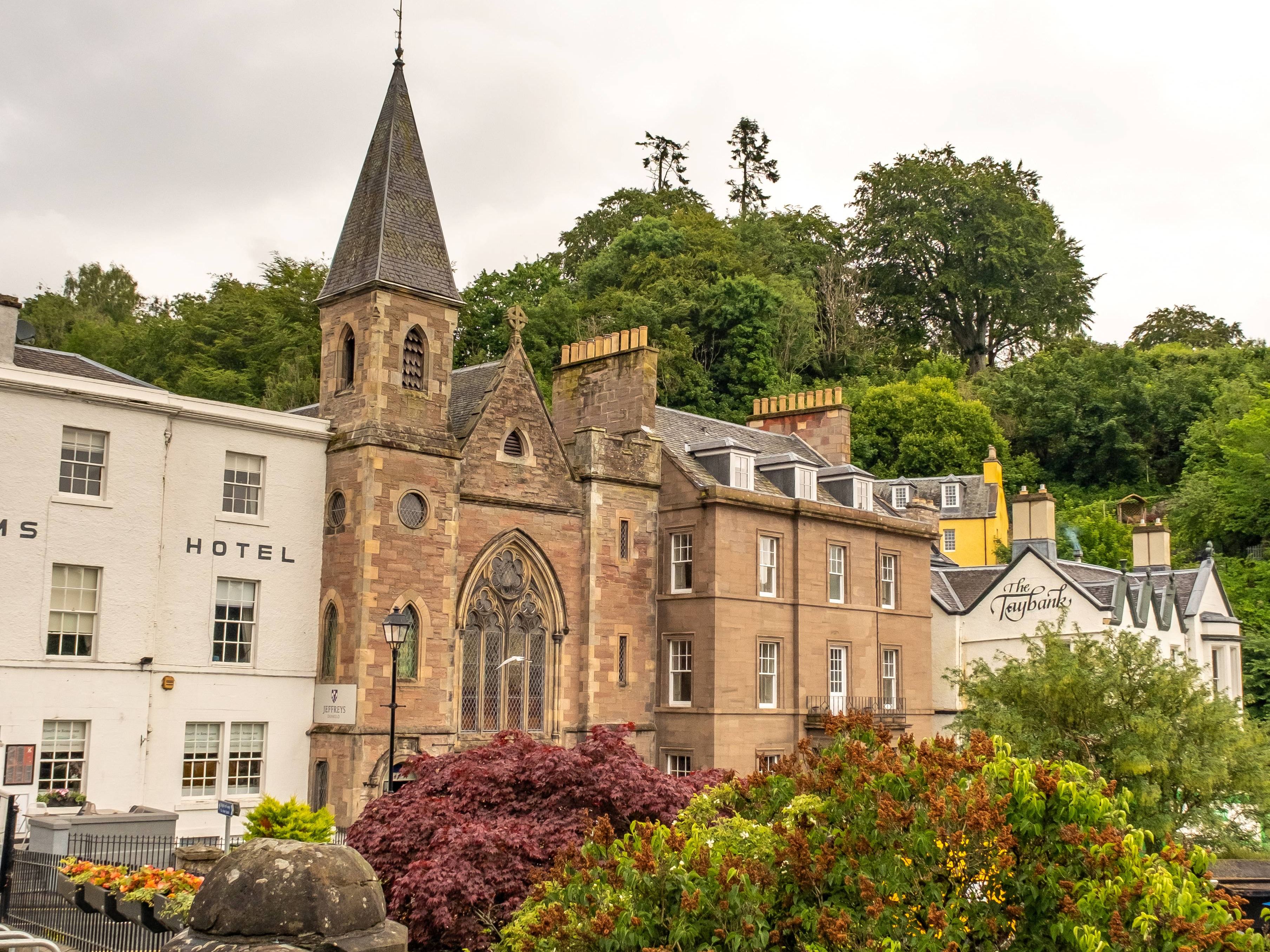 Stone church-like building with a tall spire beside a hotel, viewed over a stone wall and garden