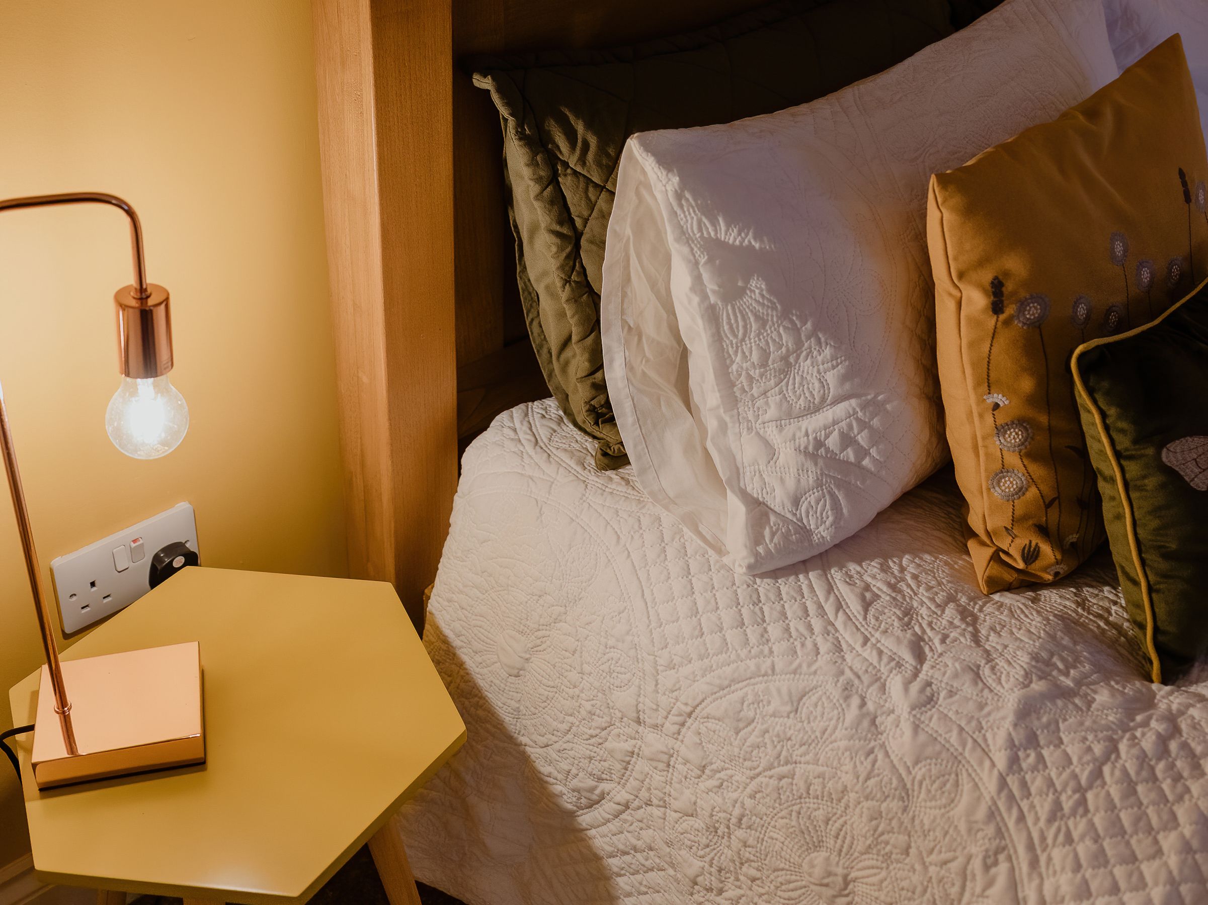 Cosy bedroom corner with wooden headboard, white quilted bedding, cushions and a bedside table lamp