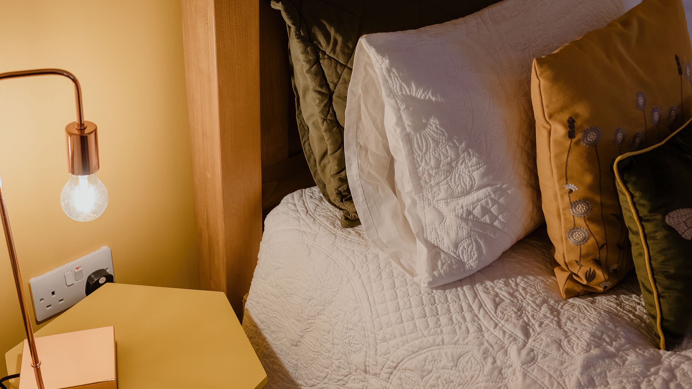 Cosy bedroom corner with wooden headboard, white quilted bedding, cushions and a bedside table lamp